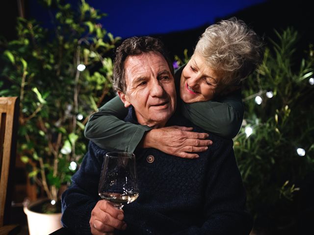 an older couple with the man sitting with a wine glass and the woman hugging from behind